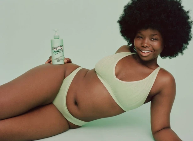 Black woman with voluminous afro hair wearing sage green underwear holding a bottle of cucumber water against a green background