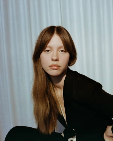 Portrait of a young woman with long brunette hair wearing a black jacket in front of a light blue draped background. Head and shoulders.