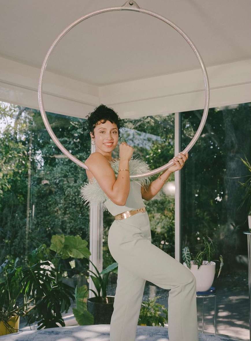 A curly-haired woman in light green outfit posing with a hoop inside a sunroom filled with plants.