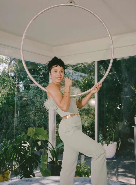 A curly-haired woman in light green outfit posing with a hoop inside a sunroom filled with plants.