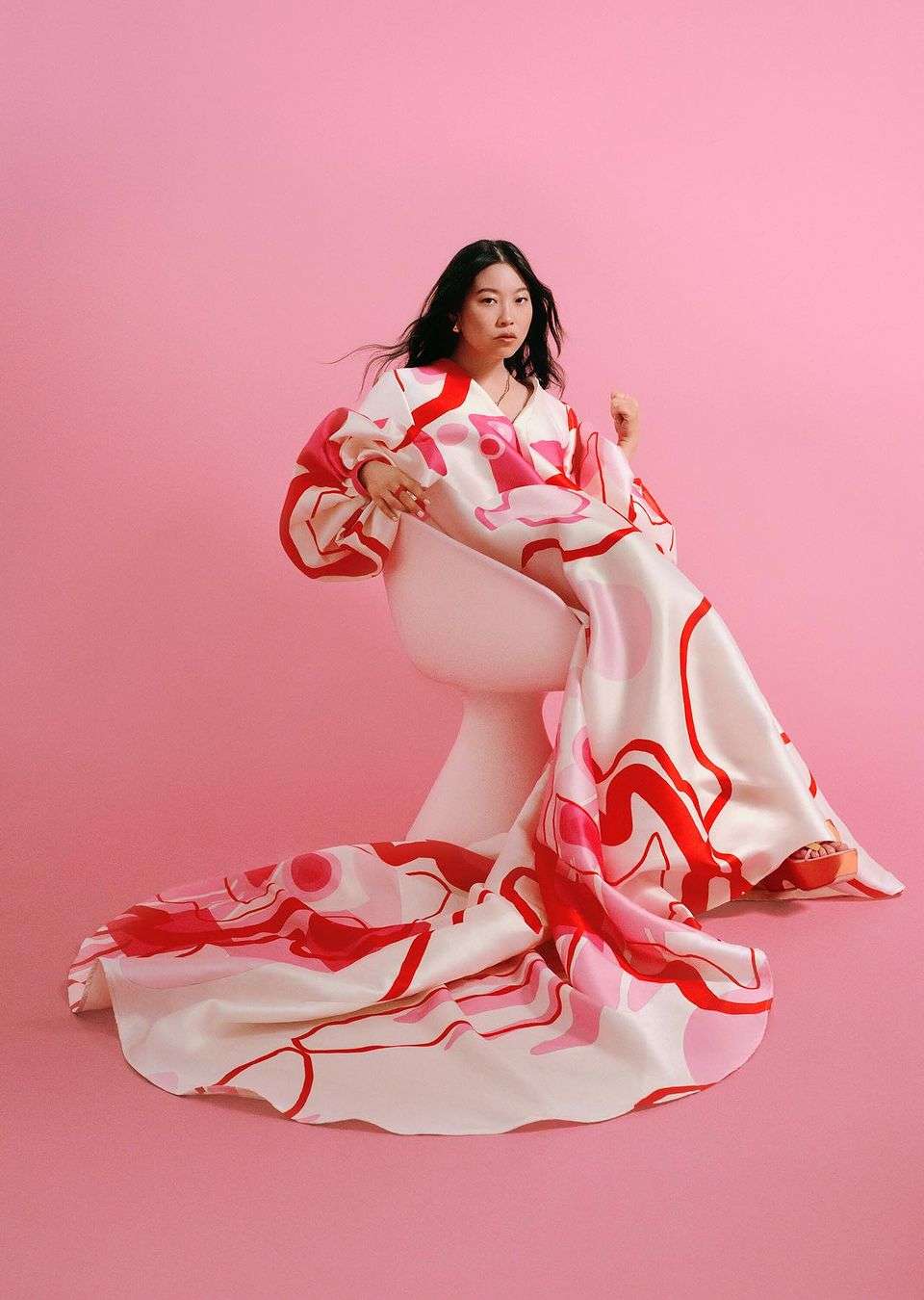 Woman in a white and pink patterned gown sits on a white chair against a pink background in a fashion photo shoot.