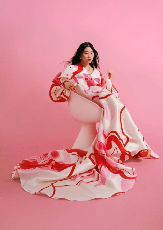 Woman in a long white dress with red abstract prints seated on a white chair against a pink backdrop