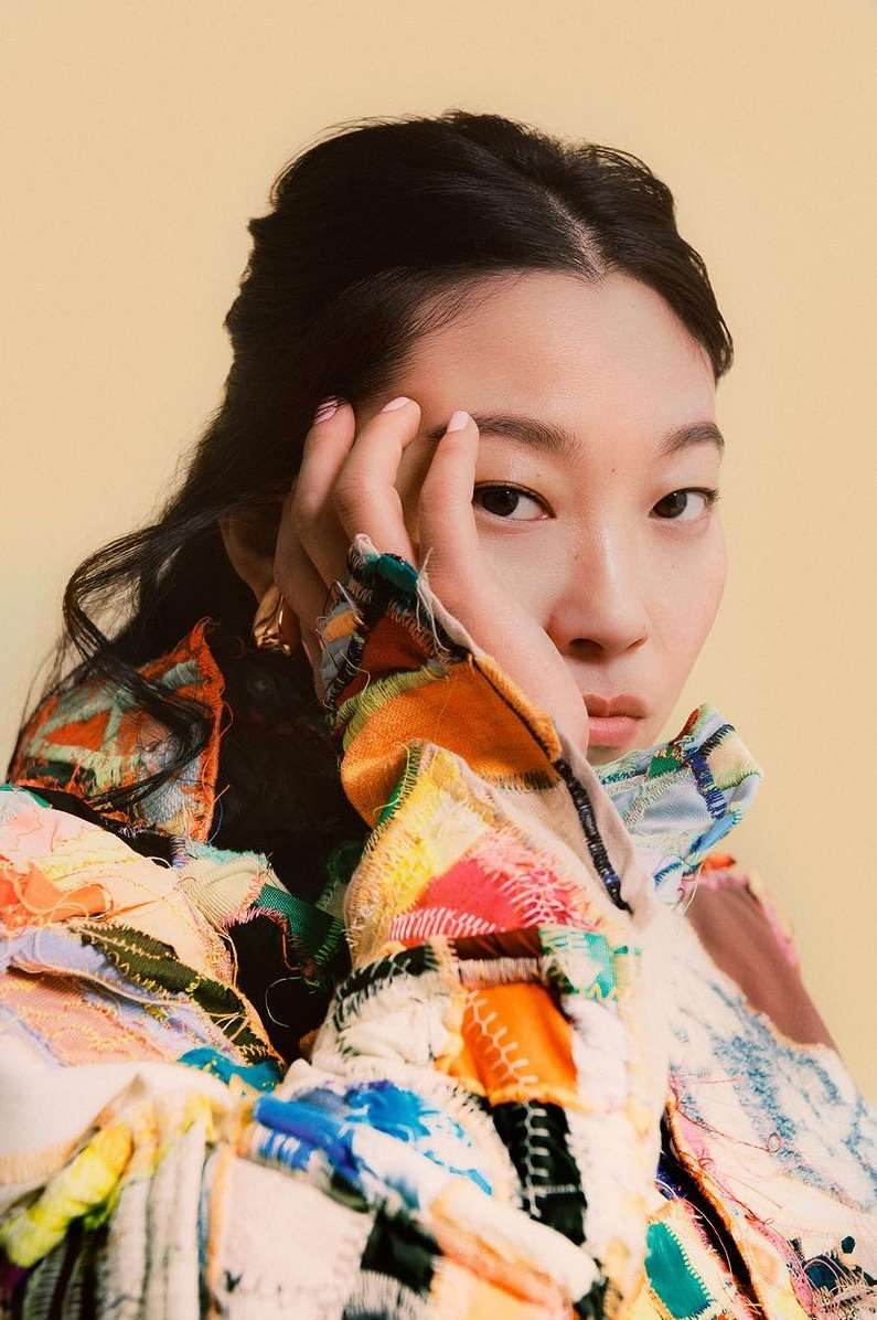 Asian female portrait in colorful patchwork jacket. Woman with dark hair poses indoors against a pale yellow backdrop, fashion.