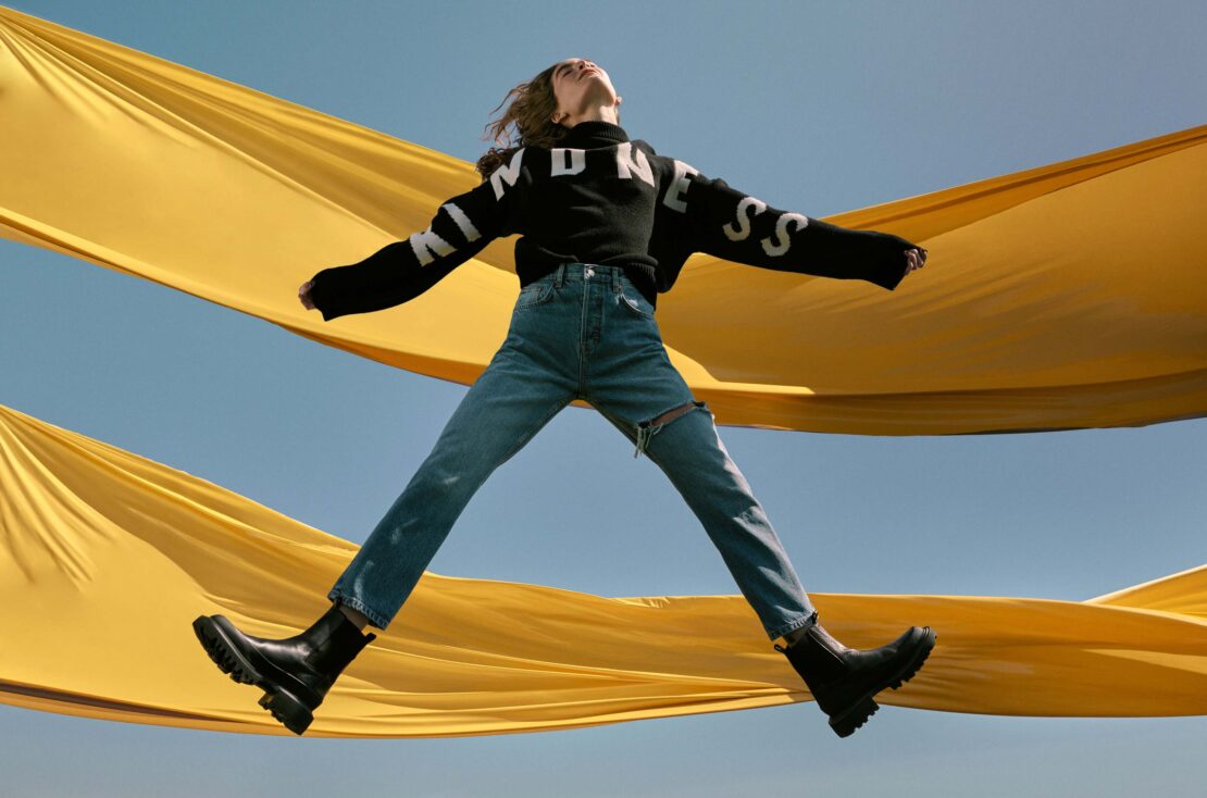 A young woman jumps in front of a yellow fabric and blue sky, wearing jeans, a black "KINDNESS" sweater, and black boots in a dynamic fashion shot.