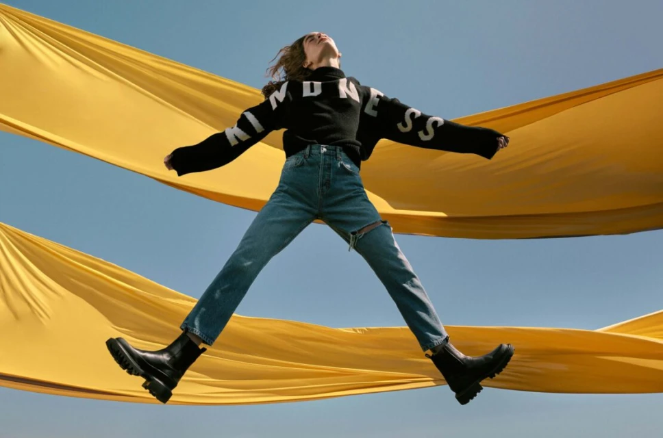 A young woman jumps in front of a yellow fabric and blue sky, wearing jeans, a black "KINDNESS" sweater, and black boots in a dynamic fashion shot.