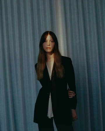 A woman in a black blazer and white shirt stands in front of vertical pleated fabric curtains with warm lighting.