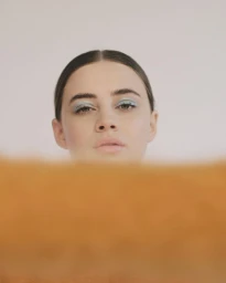 Close-up portrait of a woman with dark hair and blue eyeshadow, partially obscured by an orange foreground element against a plain background.
