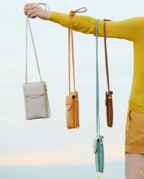Person holding up four small crossbody leather bags in different colors against a clear sky background