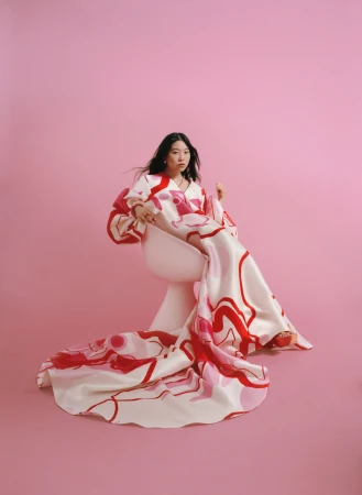 Woman wearing a patterned dress with a long train seated in a white chair against a solid pink backdrop, photographed by Amy Harrity.