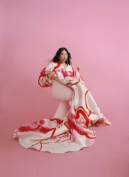 Woman wearing a patterned dress with a long train seated in a white chair against a solid pink backdrop, photographed by Amy Harrity.