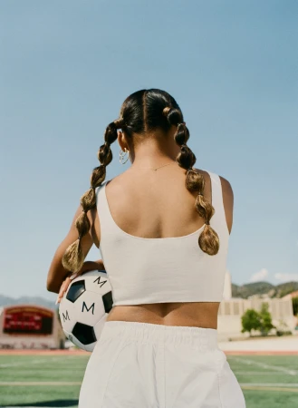 Rear view of a woman with bubble pigtails holding a soccer ball on a sports field under a clear blue sky