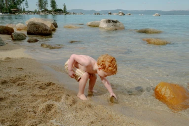 Young child with curly red hair reaching for a rock in the shallow water at a sandy beach.