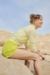 Woman in pale yellow long-sleeve top and shorts sitting outdoors on rocks under a clear blue sky, photographed by Amy Harrity.