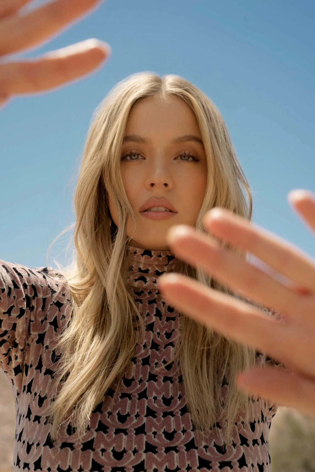 Portrait of a blonde woman wearing a patterned turtleneck top with her hands raised close to the camera lens against a clear blue sky