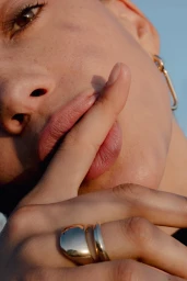 Close-up portrait of a person with a finger near lips, wearing gold statement rings and hoop earrings against clear sky.