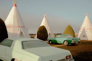 Vintage teepee-style motel buildings with retro teal and light green cars parked outside under a clear sky.