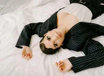 Woman in dark pinstripe blazer and white corset lying on white sheets, photographed from above by Amy Harrity.