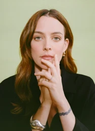 Portrait of a woman with brown hair holding her hands up near her face, wearing a black top and silver jewelry, against a green background.