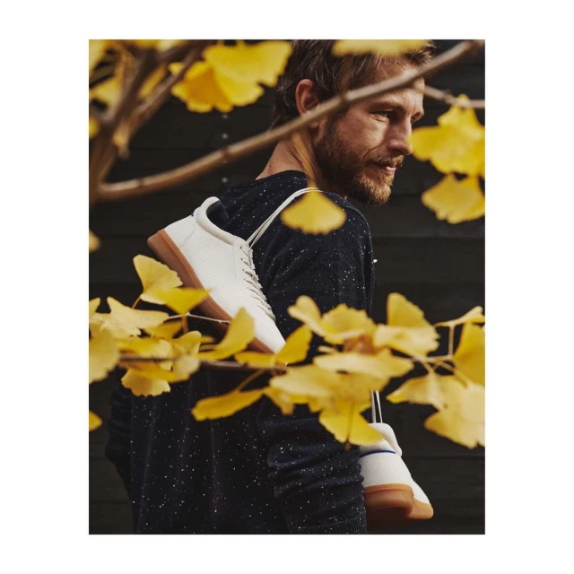 Bearded adult male wearing a sweater with white sneakers hanging around his neck outdoors, with bright yellow leaves in the foreground.