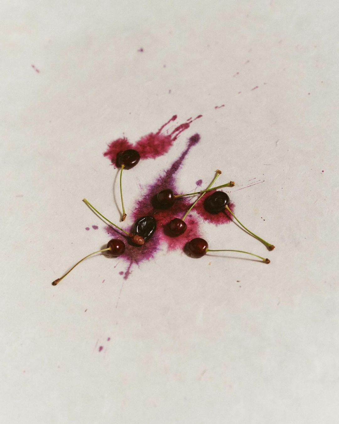 Overhead shot of cherries with stems placed on a white surface with burgundy liquid splashes, creating a minimalist food still life.