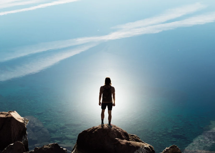 Back view of a lone adult standing on a rocky peak overlooking a calm, reflective blue lake under a bright sky with light cloud cover.