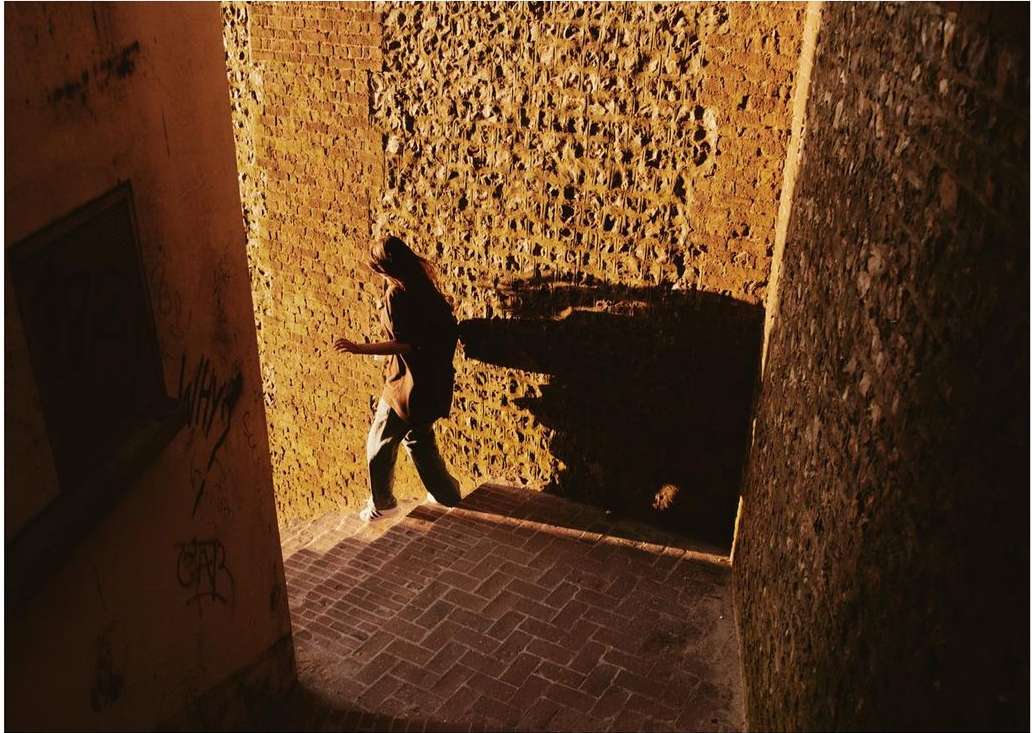 Woman in t-shirt and jeans walking down brick steps in an alleyway between two rough concrete walls in golden sunlight.