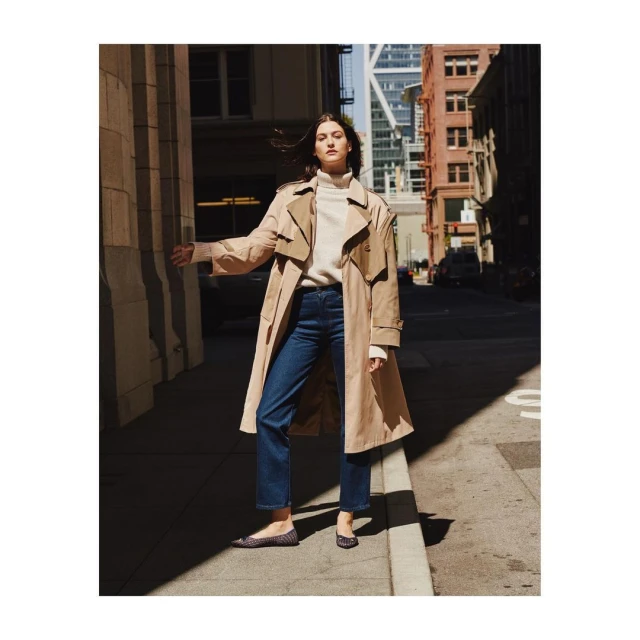 An adult woman in a trench coat and blue jeans poses in an urban alleyway between tall buildings, basking in the soft natural light.