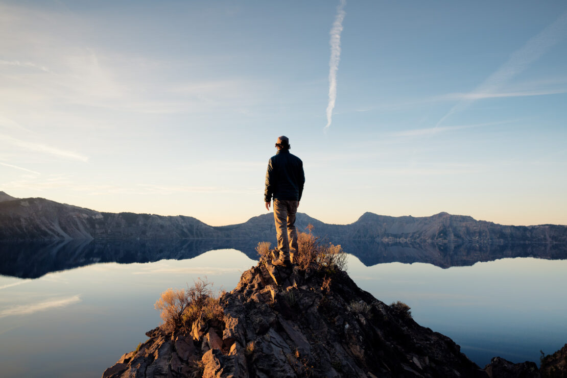 Man standing on a rocky peak overlooking a serene lake and distant mountains beneath a clear sky with a prominent contrail.