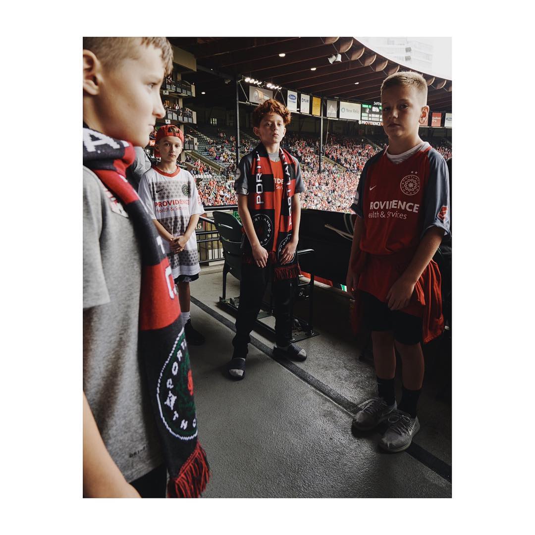 Four young soccer fans at a stadium, wearing team scarves and jerseys, with a blurred crowd in the background on a sunny day.
