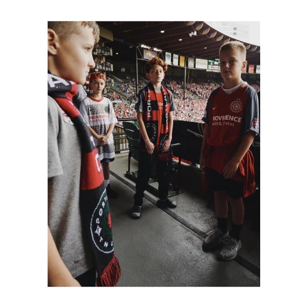 Four young soccer fans at a stadium, wearing team scarves and jerseys, with a blurred crowd in the background on a sunny day.