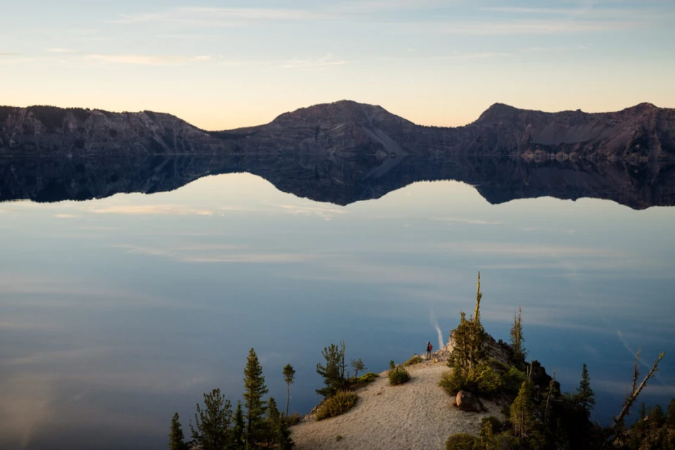 Scenic view of Crater Lake with reflections and a hiker on the edge of a ridge, surrounded by trees under a pastel sky at sunset.
