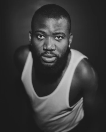 Portrait of a Black man with a beard wearing a white tank top against a dark background, photographed by Clayton Cotterell.