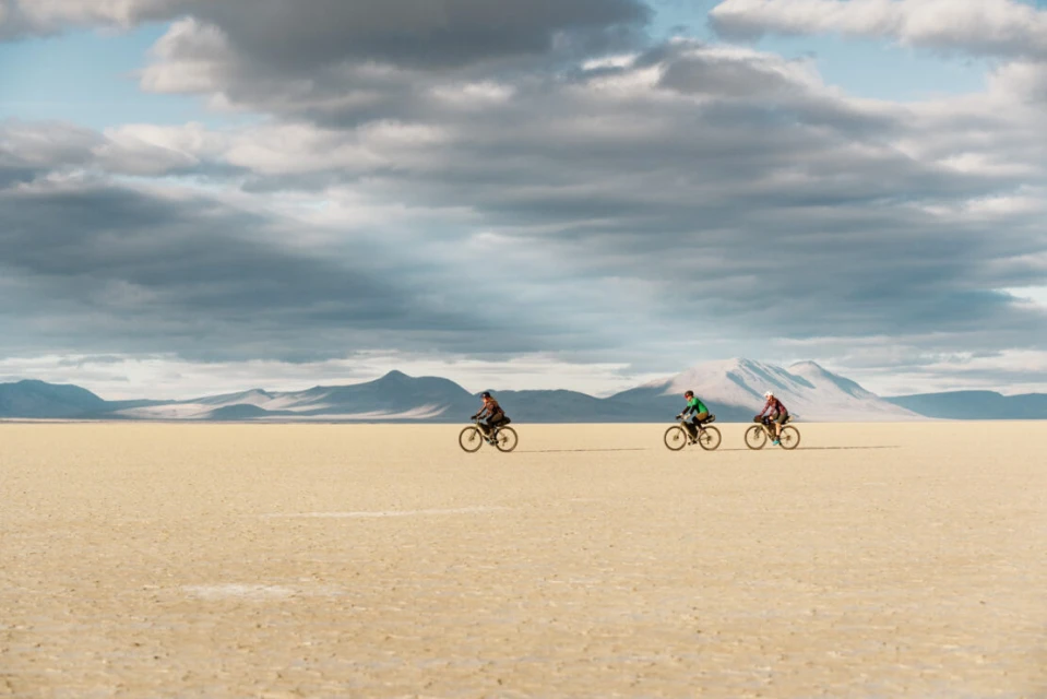 Three cyclists ride mountain bikes across a flat, dry plain with mountains in the distance under a cloudy blue sky, conveying outdoor adventure.
