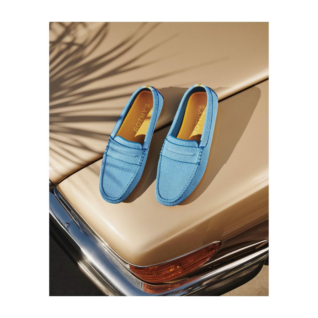 A pair of light blue Rothy's loafers sit atop the hood of a vintage beige car, with shadows from a palm frond above.