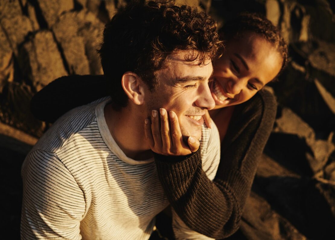 Smiling couple embracing outside in warm light. Man in striped shirt with hand on chin. Woman with dark hair hugs him, both happy.
