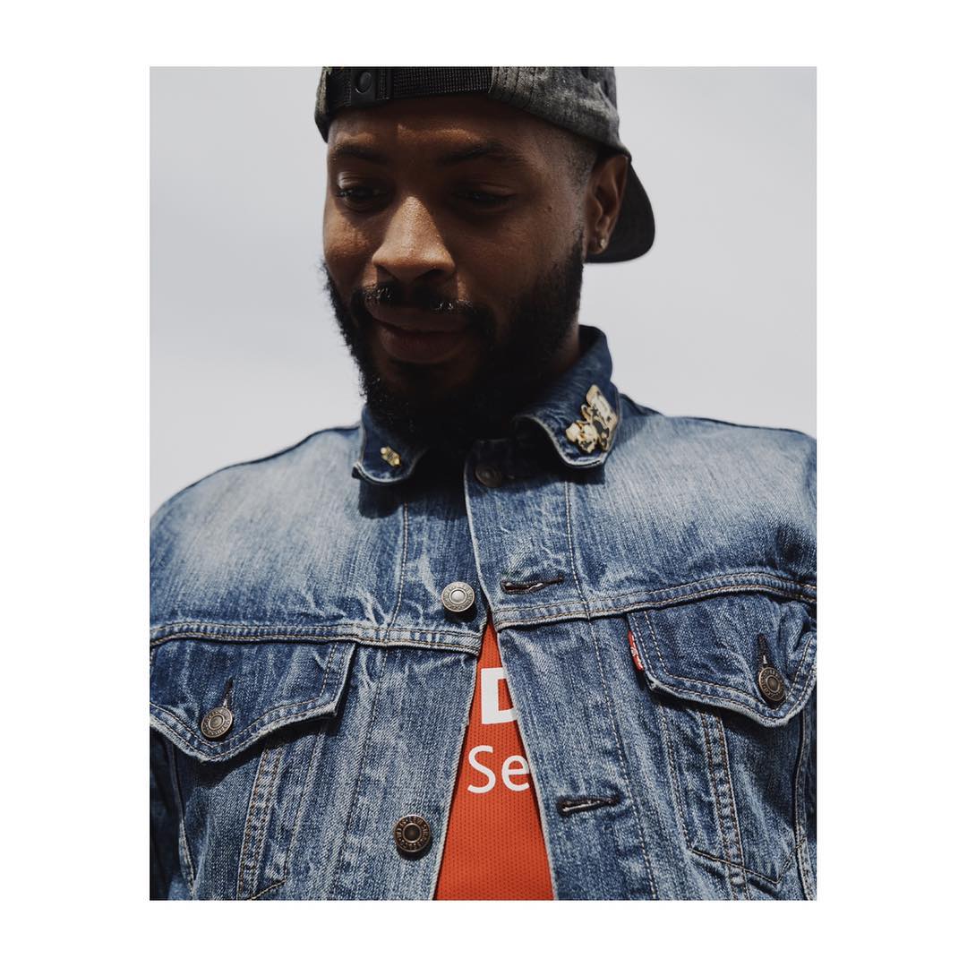 An adult male with a beard wears a denim jacket with pins, posing outdoors in a portrait with a medium shot and soft natural lighting.