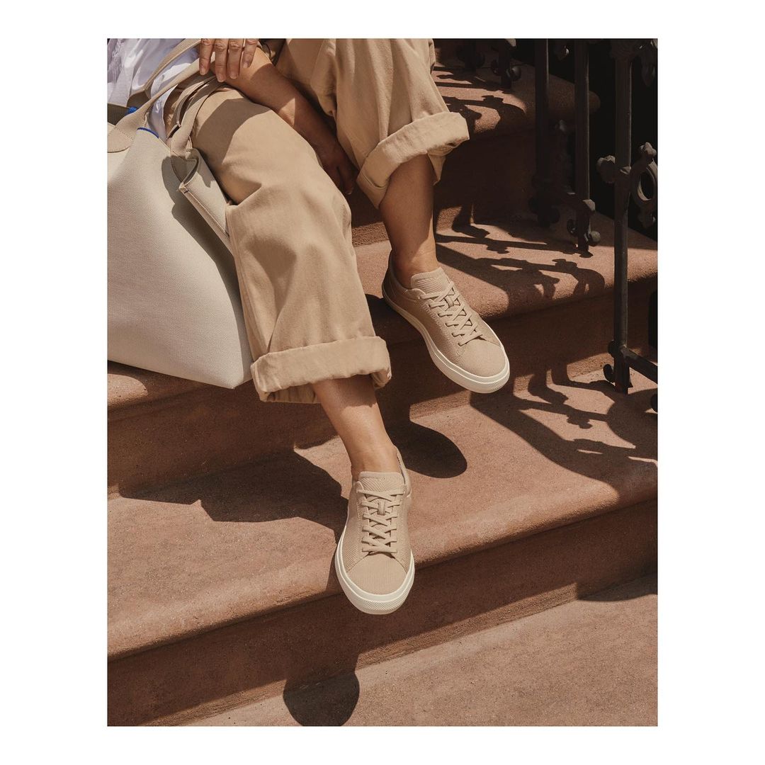 A person sitting on outdoor steps wears beige sneakers with matching cuffed pants and a shoulder bag, bathed in natural light and shadows.