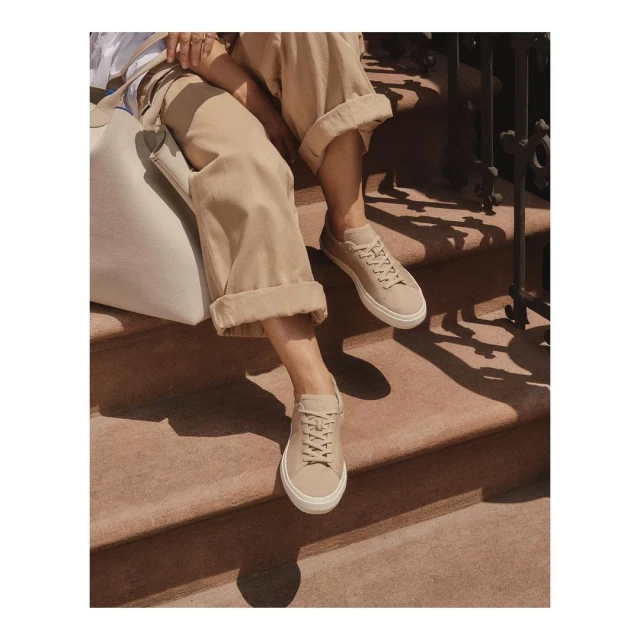 A person sitting on outdoor steps wears beige sneakers with matching cuffed pants and a shoulder bag, bathed in natural light and shadows.