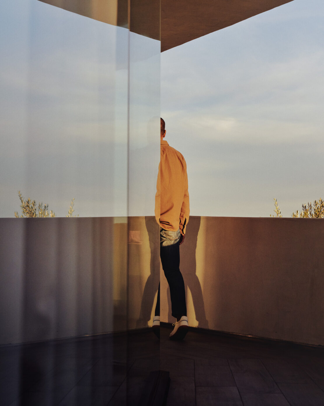Person wearing an orange shirt stands on a patio with a glass wall and partially reflects a blurred sky during the daytime.