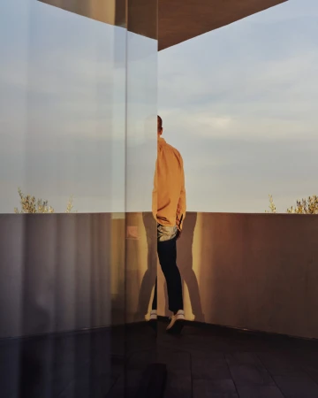 Person wearing an orange shirt stands on a patio with a glass wall and partially reflects a blurred sky during the daytime.