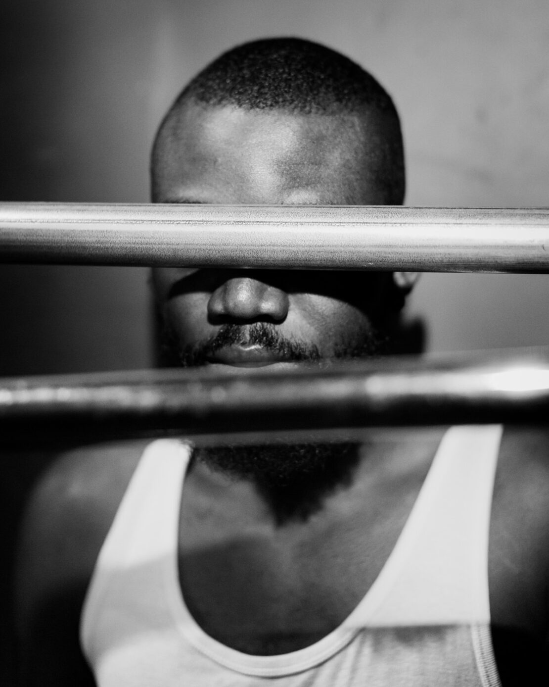 Black and white portrait of an African American adult male partially obscured by metal bars, wearing a tank top.