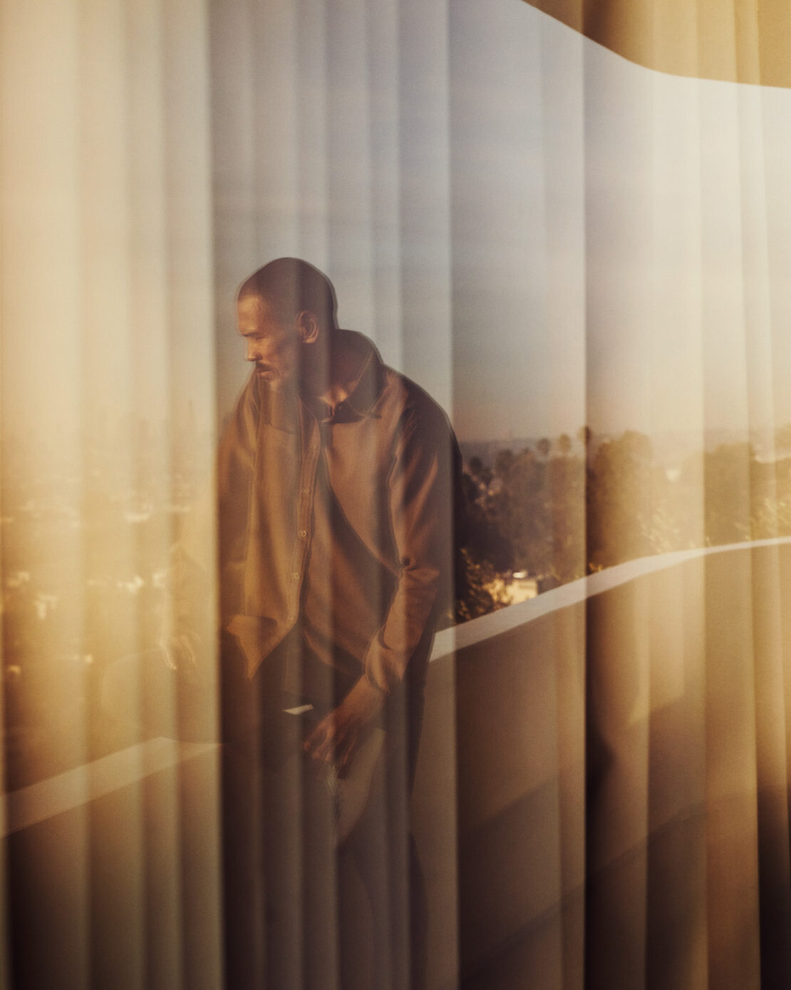 Adult male wearing a button-down shirt looking toward a city view from behind a glass enclosure with light filtering through.