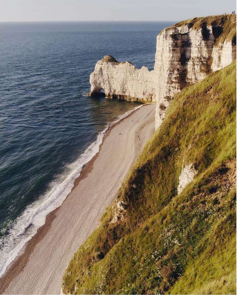 Elevated view of a sandy beach next to grass-covered cliffs and rock formations, and ocean on a sunny day, capturing the coastline.