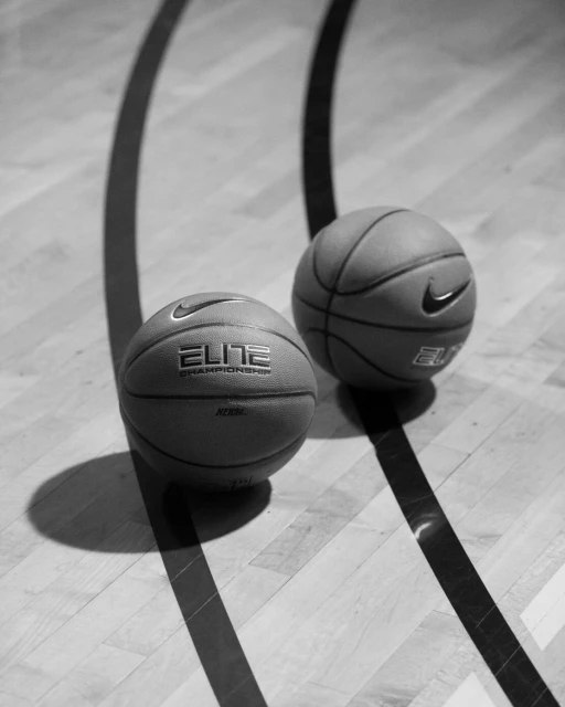 Two Nike Elite Championship basketballs rest on a wooden court with dark lines in a high-angle, black-and-white sports scene.