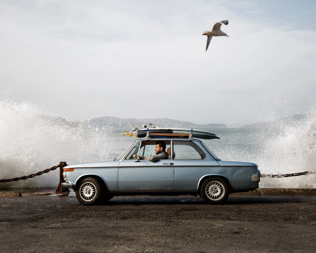 Man driving a vintage light blue car parked by the ocean with waves crashing against a concrete wall