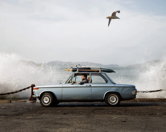 Man driving a vintage light blue car parked by the ocean with waves crashing against a concrete wall
