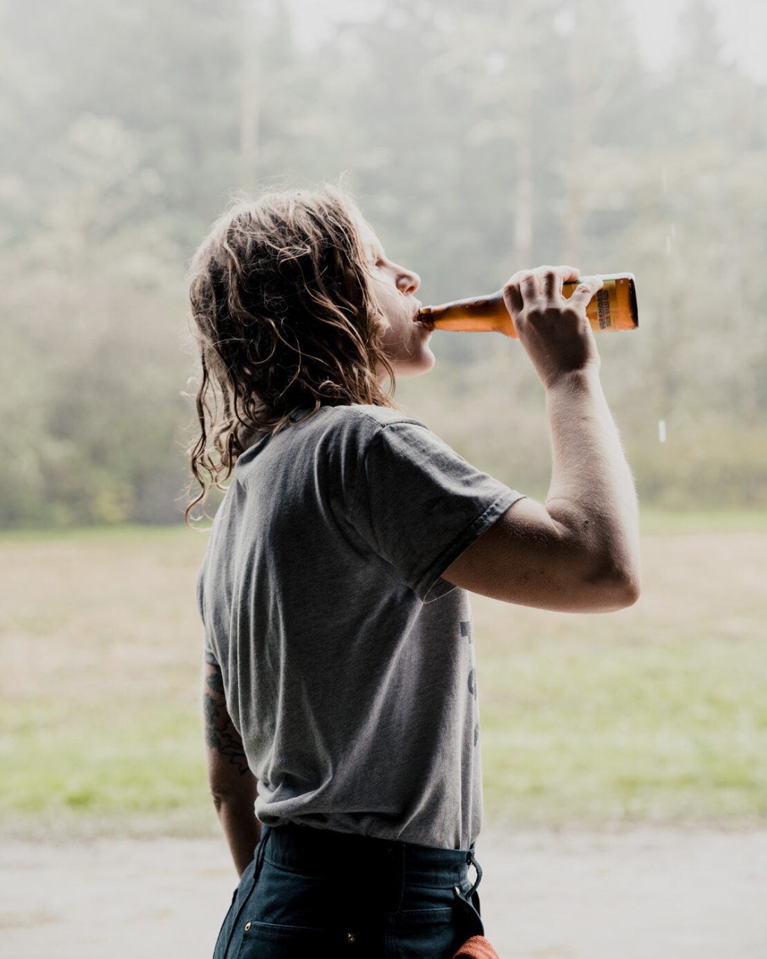 Adult woman drinking from a beer bottle, wearing a gray t-shirt and dark denim jeans in a blurred outdoor environment.