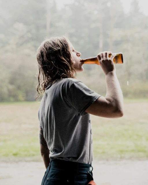 Adult woman drinking from a beer bottle, wearing a gray t-shirt and dark denim jeans in a blurred outdoor environment.