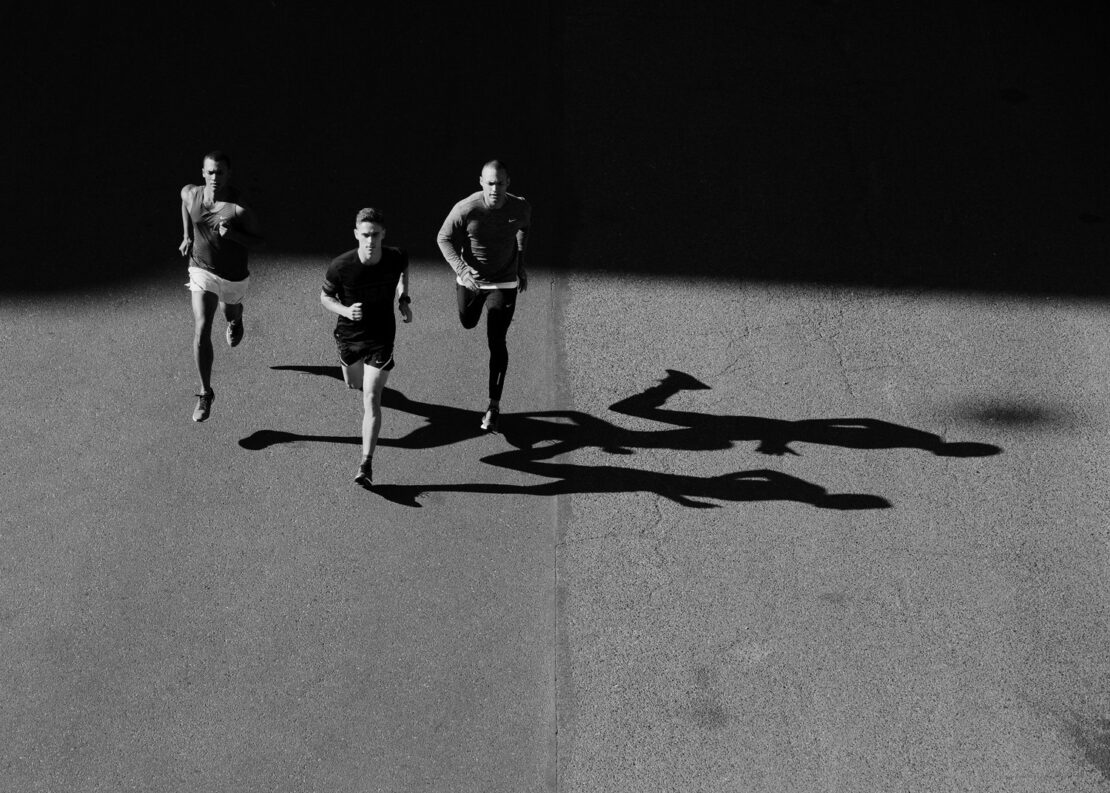 Three adult male runners are seen from an overhead perspective on an asphalt surface with long shadows in black and white.