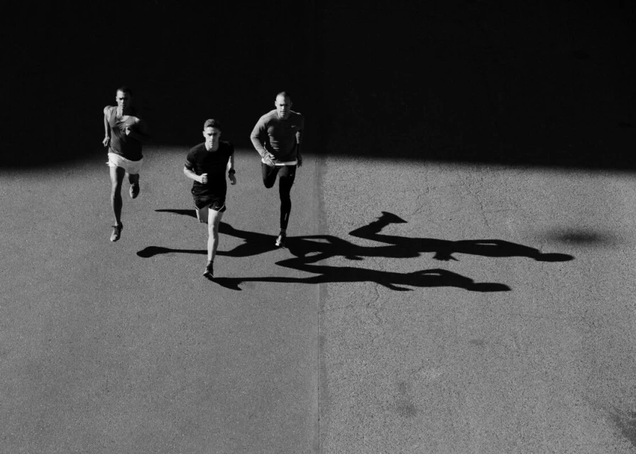 Three adult male runners are seen from an overhead perspective on an asphalt surface with long shadows in black and white.